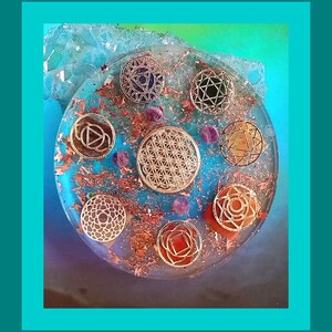 May include: A clear resin coaster with gold and copper accents features seven chakra symbols, each with a different color. The coaster also includes a flower of life symbol in the center.