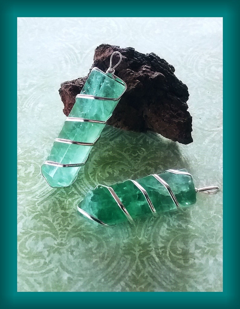 May include: Two green fluorite crystal pendants wrapped in silver wire. The crystals are a light green colour and have a natural, rough texture. The wire is wrapped around the crystals in a spiral pattern.