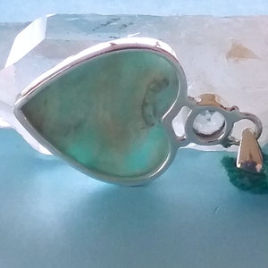 May include: A silver heart-shaped pendant with a light green stone set in the centre. The pendant has a small clear stone on the side and a loop for attaching a chain.