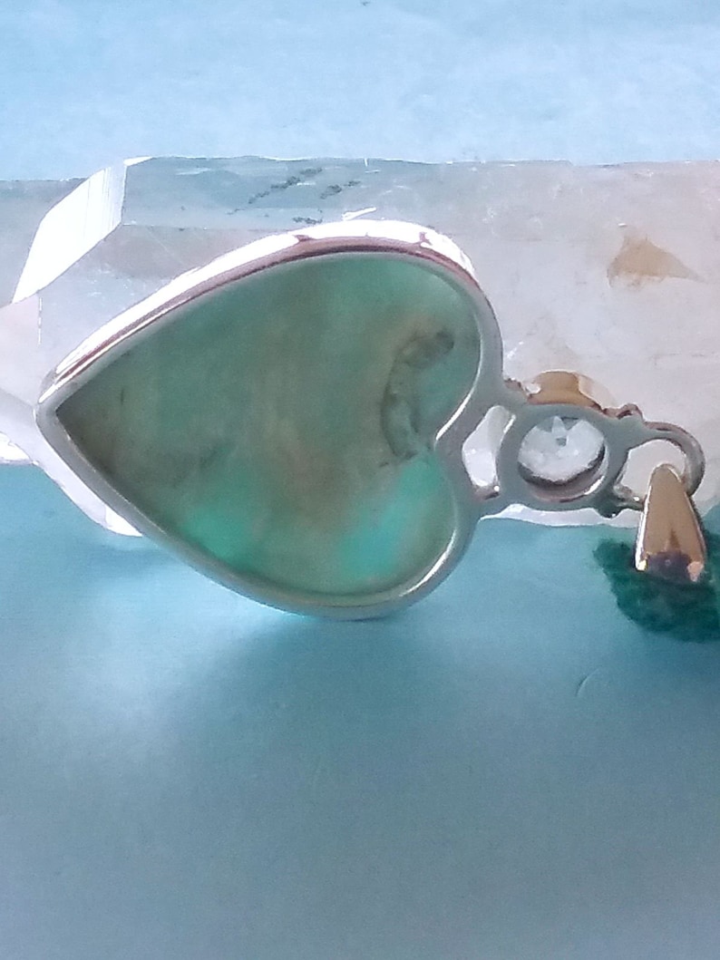 May include: A silver heart-shaped pendant with a light green stone set in the centre. The pendant has a small clear stone on the side and a loop for attaching a chain.