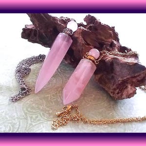 May include: Two pink rose quartz crystal pendants with silver and gold chains. One is a long, pointed shape, the other rectangular. Both have a small, round crystal at the top. The pendants are displayed on a light green surface.