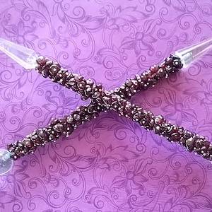 Może przedstawiać: Para czarodziejskich różdżek wykonanych z ciemnoczerwonych koralików z kamieni szlachetnych i przezroczystych kryształowych końcówek. Różdżki są skrzyżowane w kształcie X.