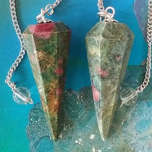 May include: Two green and red gemstone pendulums with silver chains and clear crystal beads. The pendulums are pointed and have a faceted design.