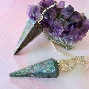 Könnte beinhalten: Zwei facettierte, kegelförmige Anhänger aus dunkelgrünem und blaugrünem Mineralgestein, jeweils an einer silberfarbenen Metallkette befestigt. Ein Anhänger liegt an einer großen, gezackten Ansammlung tiefvioletter Amethystkristalle mit durchscheinenden Kanten.