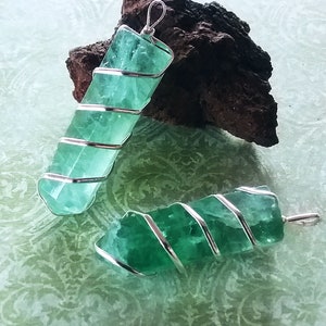 May include: Two green fluorite crystal pendants wrapped in silver wire. The pendants are on a green patterned surface.