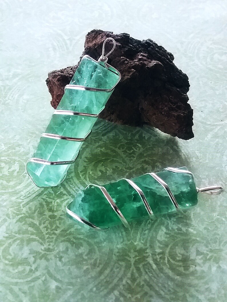 May include: Two green fluorite crystal pendants wrapped in silver wire. The pendants are on a green patterned surface.