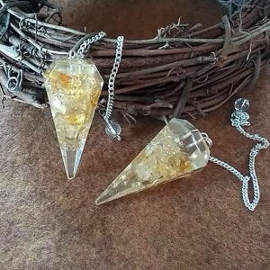 May include: Two gold and clear crystal point pendulums with silver chains. The pendulums are hanging from a brown woven wreath.