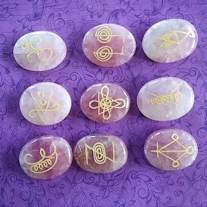 May include: Nine pink oval-shaped stones with gold symbols etched on them. The stones are arranged in three rows of three.