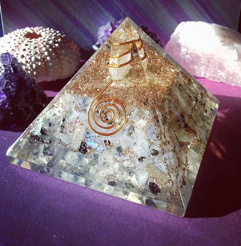 May include: A clear resin pyramid containing embedded white and translucent stone fragments, gold-colored metallic flakes, and a central clear quartz point wrapped in a copper wire coil, featuring a prominent copper spiral on the front face.