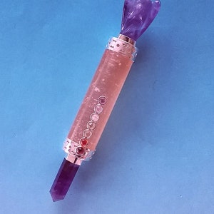 May include: A purple amethyst crystal wand with an angel figure at the top and a pointed crystal at the bottom. The wand has a clear, cylindrical body with embedded gemstones and silver-coloured metal accents. The background is a solid blue.