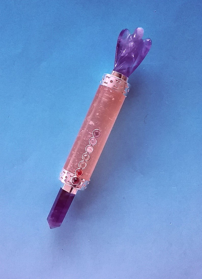 May include: A purple amethyst crystal wand with an angel figure at the top and a pointed crystal at the bottom. The wand has a clear, cylindrical body with embedded gemstones and silver-coloured metal accents. The background is a solid blue.