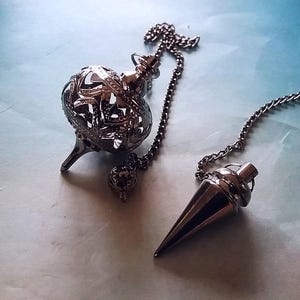 May include: Two metal pendulums on a light blue surface. One pendulum has a chain and a decorative, openwork sphere. The other pendulum is cone-shaped with a chain. Both are silver in color.
