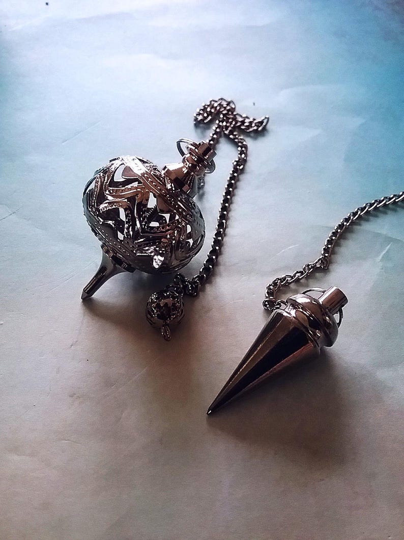May include: Two metal pendulums on a light blue surface. One pendulum has a chain and a decorative, openwork sphere. The other pendulum is cone-shaped with a chain. Both are silver in color.