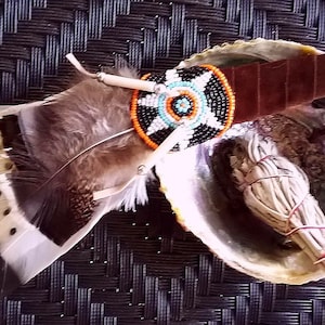 May include: A Native American smudging kit with a decorated feather, a beaded fan, a bundle of sage, and a suede leather strap. The kit is arranged in an abalone shell, on a dark woven background.