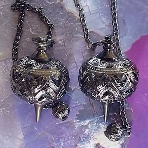 May include: Two ornate, dark metal pendulums with intricate filigree designs and pointed tips hang from chains. Each pendulum has a small, decorative sphere attached to the chain below the main body. The background is a blurred mix of purple and blue.
