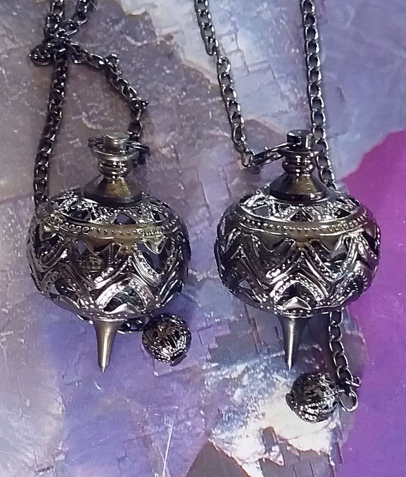May include: Two ornate, dark metal pendulums with intricate filigree designs and pointed tips hang from chains. Each pendulum has a small, decorative sphere attached to the chain below the main body. The background is a blurred mix of purple and blue.