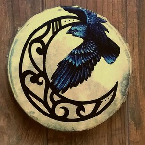 May include: A circular wooden plaque featuring a hand-painted illustration of a dark blue raven in flight positioned in front of a large, black, stylized crescent moon. The background of the moon is a pale, mottled cream and light green color.