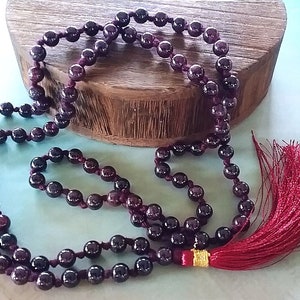 May include: A dark red gemstone mala necklace with a red tassel. The necklace is made of round beads and is strung on a dark red cord.