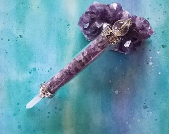 Puede incluir: Un tubo de vidrio transparente lleno de cristales de amatista, coronado con una tapa de metal plateado y dorado. El tubo descansa sobre un gran cúmulo de cristales de amatista.