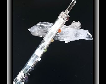 May include: A clear quartz crystal wand with a silver tip and a clear quartz crystal sphere at the bottom. The wand is decorated with seven small colored stones representing the seven chakras. The wand is resting on two clear quartz crystal points.