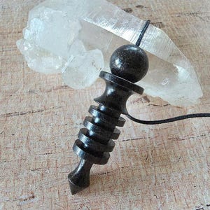 May include: A dark brown wooden pendulum featuring a spherical top, a series of four graduated circular ridges along the central shaft, and a pointed base. A thin black cord is attached to the top of the pendulum, which is positioned in front of a clear, translucent quartz crystal point.
