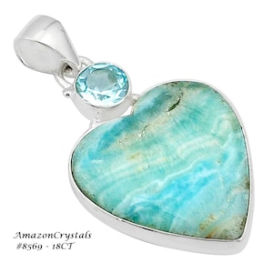 May include: A silver heart-shaped pendant with a blue gemstone set in the centre. The pendant is accented with a smaller blue gemstone at the top. The pendant is labelled "AmazonCrystals #8569 - 18CT".