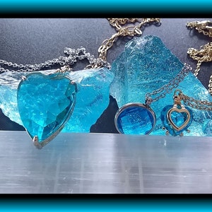 May include: Two necklaces with blue heart-shaped pendants and a blue round pendant. The necklaces are on a white surface with blue crystal rocks.