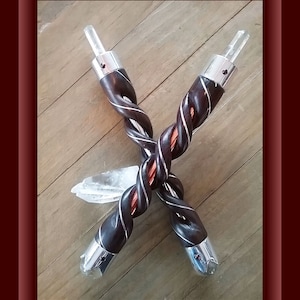 May include: Two wooden wands with a twisted design, each topped with a clear quartz crystal and a silver metal cap with a red gemstone accent. The wands are crossed over each other and a clear quartz crystal is lying on the wooden surface.