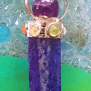 Puede incluir: Un colgante de plata con una piedra de amatista p&uacute;rpura en el centro, rodeada por un c&iacute;rculo de plata. El colgante est&aacute; unido a un cord&oacute;n negro y tiene tres piedras m&aacute;s peque&ntilde;as: naranja, amarilla y verde. El colgante cuelga sobre un fondo verde y azul.