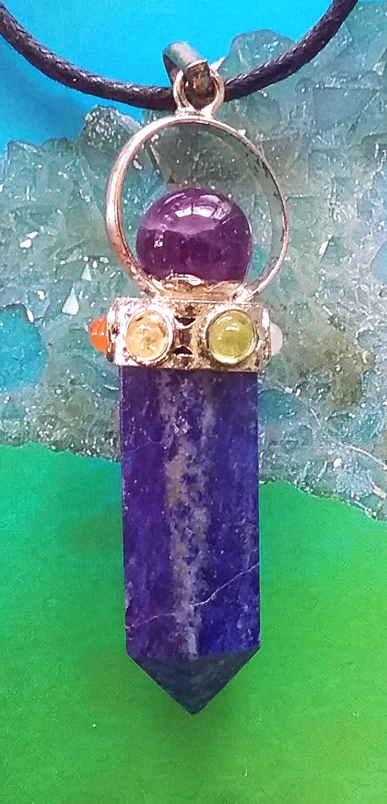 Puede incluir: Un colgante de plata con una piedra de amatista p&uacute;rpura en el centro, rodeada por un c&iacute;rculo de plata. El colgante est&aacute; unido a un cord&oacute;n negro y tiene tres piedras m&aacute;s peque&ntilde;as: naranja, amarilla y verde. El colgante cuelga sobre un fondo verde y azul.