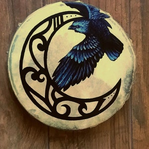 May include: A circular drum featuring a hand-painted design of a black raven in flight against a pale yellow crescent moon. The moon is decorated with black filigree patterns. The drum head has a textured, off-white surface with visible edges.