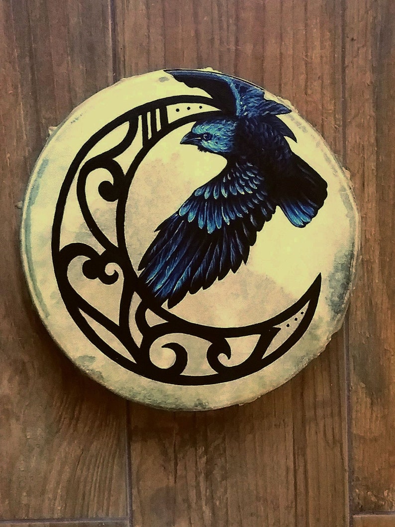 May include: A circular drum featuring a hand-painted design of a black raven in flight against a pale yellow crescent moon. The moon is decorated with black filigree patterns. The drum head has a textured, off-white surface with visible edges.