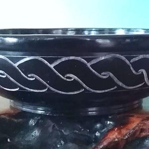 May include: A black stone bowl with a carved white design. The bowl is round and has a wide rim. It is sitting on a wooden surface.