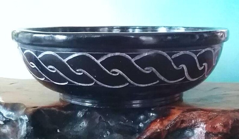 May include: A black stone bowl with a carved white design. The bowl is round and has a wide rim. It is sitting on a wooden surface.