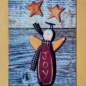 May include: A decorative pin set featuring a wooden angel with the word "JOY" and two star-shaped pins. The angel has wings and a checkered scarf. The background is a rustic wood pattern, with a tan border decorated with stars and hearts.