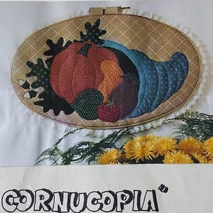 May include: A framed fabric art piece featuring a cornucopia design. The artwork includes a variety of colorful fruits and vegetables, with the text "CORNUCOPIA" and dimensions of 18" x 27". The Osage County Quilt Factory presents this piece.