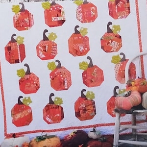 May include: A quilt pattern titled Sewn into the fabric... Pieces of our lives and Patch Work 58" x 70". The quilt features a white background with a repeating pattern of orange pumpkins with green stems. A wooden chair sits nearby with fabric pumpkins.