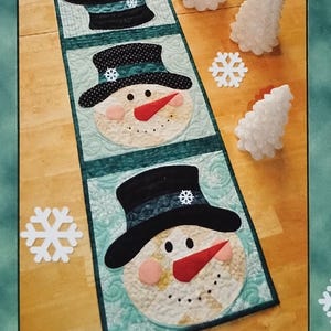 May include: A rectangular patchwork table runner featuring four snowman faces wearing black top hats. The snowmen have orange carrot noses, black eyes, and pink cheeks. The runner is set against a light blue background with white snowflake cutouts.