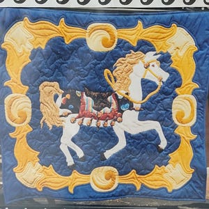 May include: A blue and gold carousel pony quilt pattern by Shirley Gardner Designs. The quilt features a white pony with a flowing mane and tail, a colorful saddle, and a gold frame. The text "The Carousel Pony" is also visible.