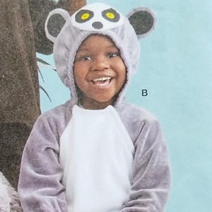 May include: A child wearing a gray lemur costume with a white chest, black mittens, and a striped tail. The costume includes a hood with lemur ears and facial details. The child is smiling.