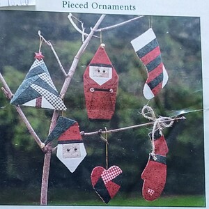 May include: A kit for making six patchwork Christmas ornaments. The kit includes fabrics, muslin, embellishments, and instructions. The ornaments include a Christmas tree, Santa, a stocking, a mitten, and a heart. The kit is packaged in a clear plastic bag.