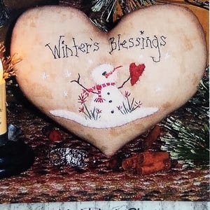 May include: A heart-shaped fabric ornament with a tan, tea-stained appearance. The text "Winter's Blessings" is printed above a small snowman illustration wearing a red and white striped scarf and holding a red heart. The border features stars.