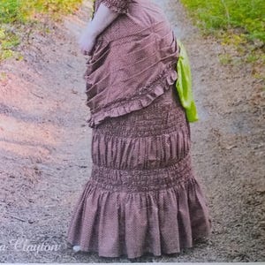 May include: A McCall's Costumes sewing pattern envelope M8191 featuring a brown patterned dress with a ruffled skirt, three-quarter sleeves, and a bright green accent piece. The text includes McCall's, COSTUMES, M8191, SIZE A5 (6-8-10-12-14), and Angela Clayton.