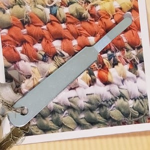 May include: A metal tool for making rag rugs, featuring a flat handle with a hook at one end and a hole at the other with a ribbon attached. The tool is displayed on a card titled RAG RUGS with Easy, Detailed Rectangular Rug Instructions.