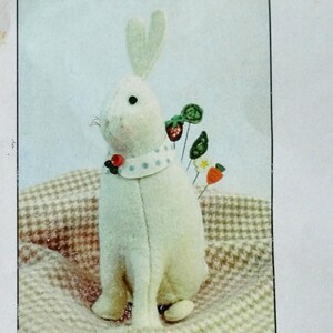 May include: A cream-colored Beatrix Pincushion bunny with a white collar and decorative pins. The image is on a cream and tan checkered background. The text "Beatrix Pincushion" is printed above the image. The text "Wool, roving, buttons and pins included. You add needle and thread" is printed below the image.