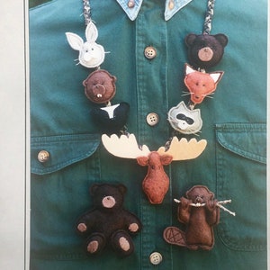 May include: A necklace of felt woodland animal figures, including a bear, rabbit, skunk, moose, raccoon, fox, and beaver. The animals are in various colors and sizes, with detailed features. The necklace is displayed on a green shirt.