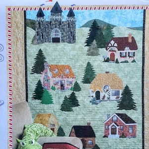 May include: A quilt pattern titled "Once Upon a Time" featuring a patchwork design of a castle, houses, and trees. The quilt is displayed with a green dragon stuffed animal and a smaller quilt with a castle design.