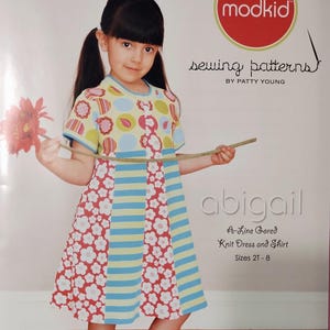May include: A young girl models a colorful A-line gored knit dress and shirt sewing pattern. The dress features a floral and striped design with the text "abigail" and "modkid" on the pattern cover. Sizes 2T-8.