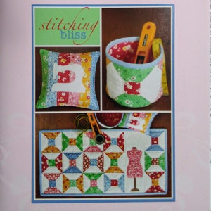 May include: The Janelle Wind Collection features a patchwork sewing-themed design. The image showcases a pillow with a sewing machine applique, a fabric basket with sewing tools, and a quilted mat with a dress form. The text "stitching bliss" is also visible.