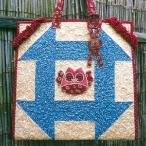May include: A quilted tote bag with a blue and cream geometric design, featuring a red owl applique. The bag has red handles and trim. The background includes a bamboo fence and greenery. Text on the bag reads "OW. Block Keeper" and "Lush the Happy Quilter".